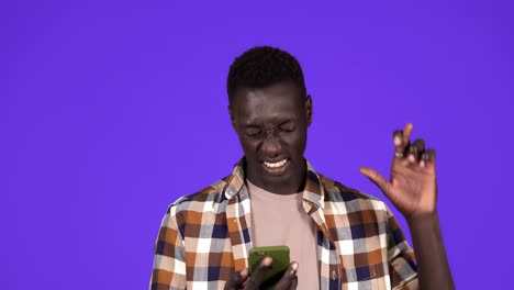 Young-black-man-holding-a-phone-crossing-fingers-for-having-luck,-looking-on-mobile-screen-and-burst-in-smiling,-winner