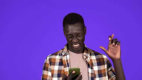 young black man holding a phone crossing fingers for having luck, looking on mobile screen and burst in smiling, winner