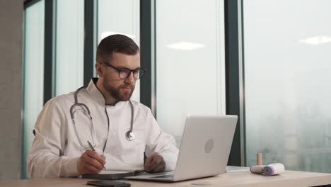 a solid male doctor sitting at a table with a laptop works and writes in a notebook