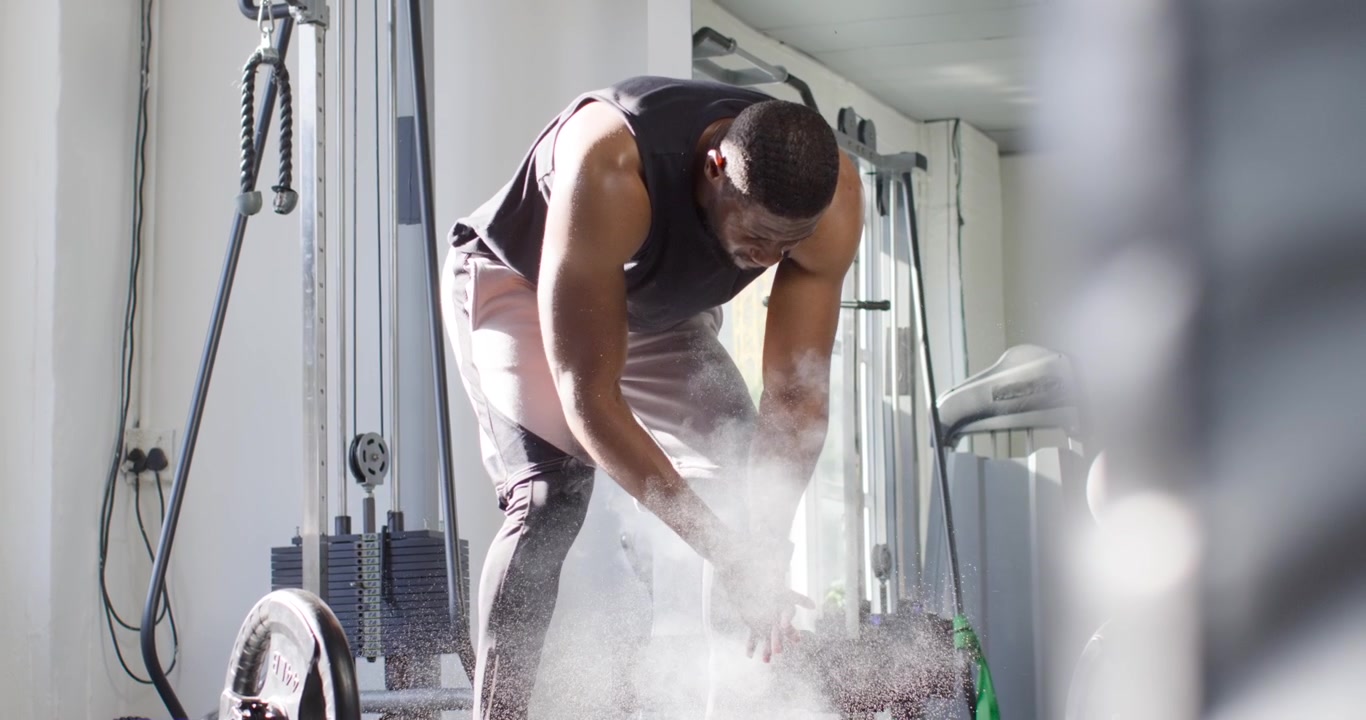 Uomo africano in canotta che si prepara per il sollevamento, si strofina il gesso e afferra il bilanciere in palestra per una migliore presa