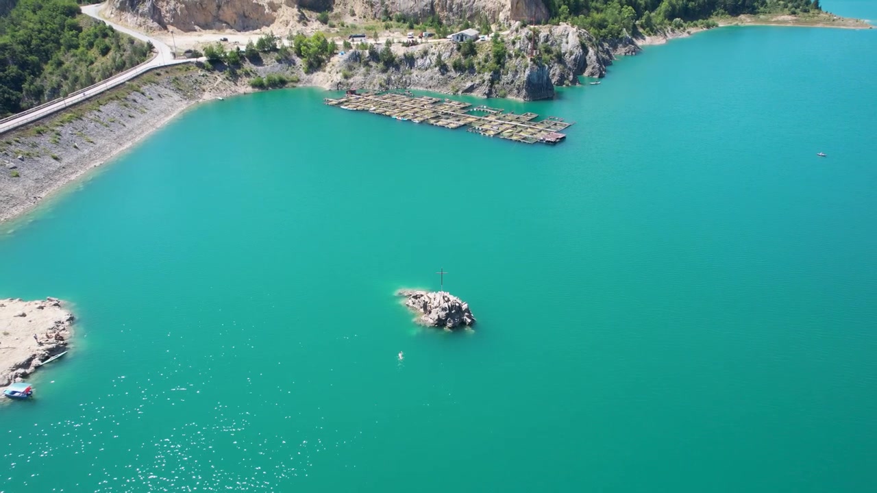 En luftdrone passerer en klippeø mod flydende fiskebure på Zaovine-søen i Serbien, med turkis vand, en stenbrudskyst, bugtende vej og skovklædte skråninger i baggrunden