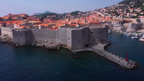 Revelación-Aérea-Volando-Hacia-Atrás-Desde-El-Puerto-Y-Las-Murallas-De-La-Ciudad-Vieja-De-Dubrovnik,-Croacia-Con-Montañas-Y-Mar-En-4k