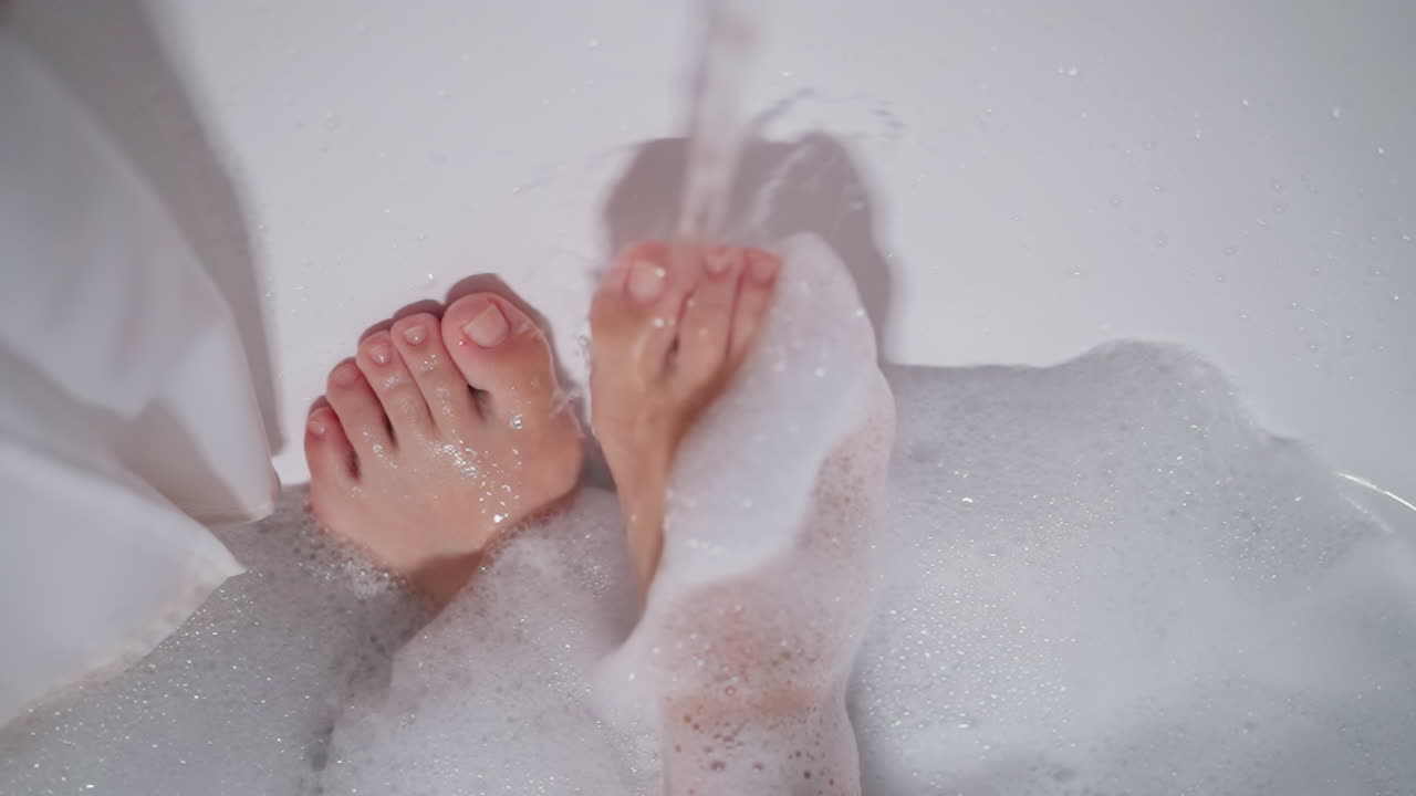 Woman washes foamy foot with water in bathtub closeup. Lady takes care of legs bathing in hot water in cozy bathroom. Relaxation time and spa