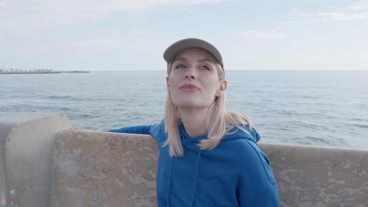 A blonde woman gazes out at the sea while seated on a stone bench along the coastline