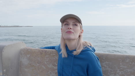 a blonde woman gazes out at the sea while seated on a stone bench along the coastline