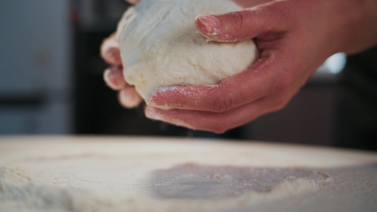 Hands knead white dough on floured surface. Motion shows dough forming, texture, and flour dust. Process captures rustic, homemade baking. Ideal for cooking, food, or lifestyle projects