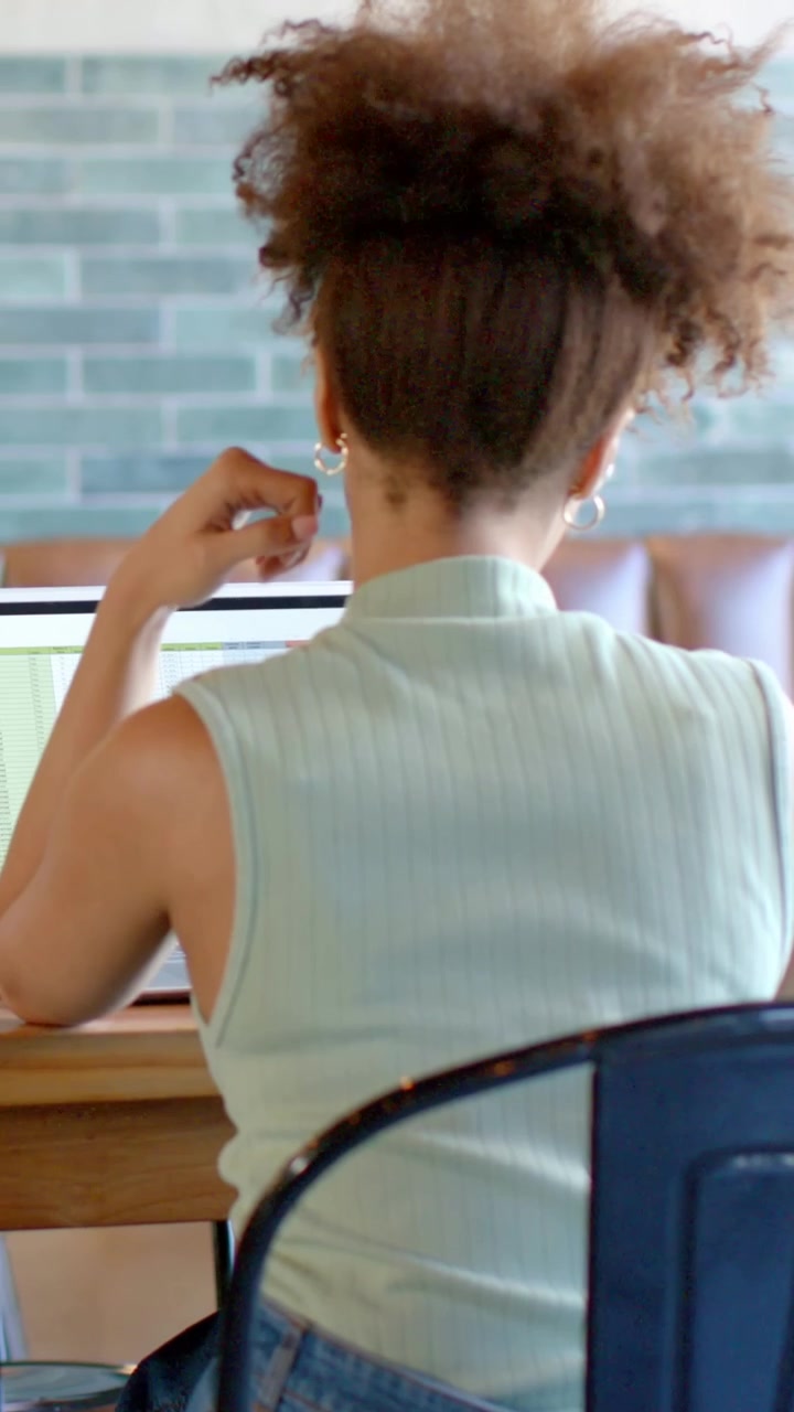 Vertical video: Working on laptop woman in teal top and jeans rubbing neck fixing hoops at cafe