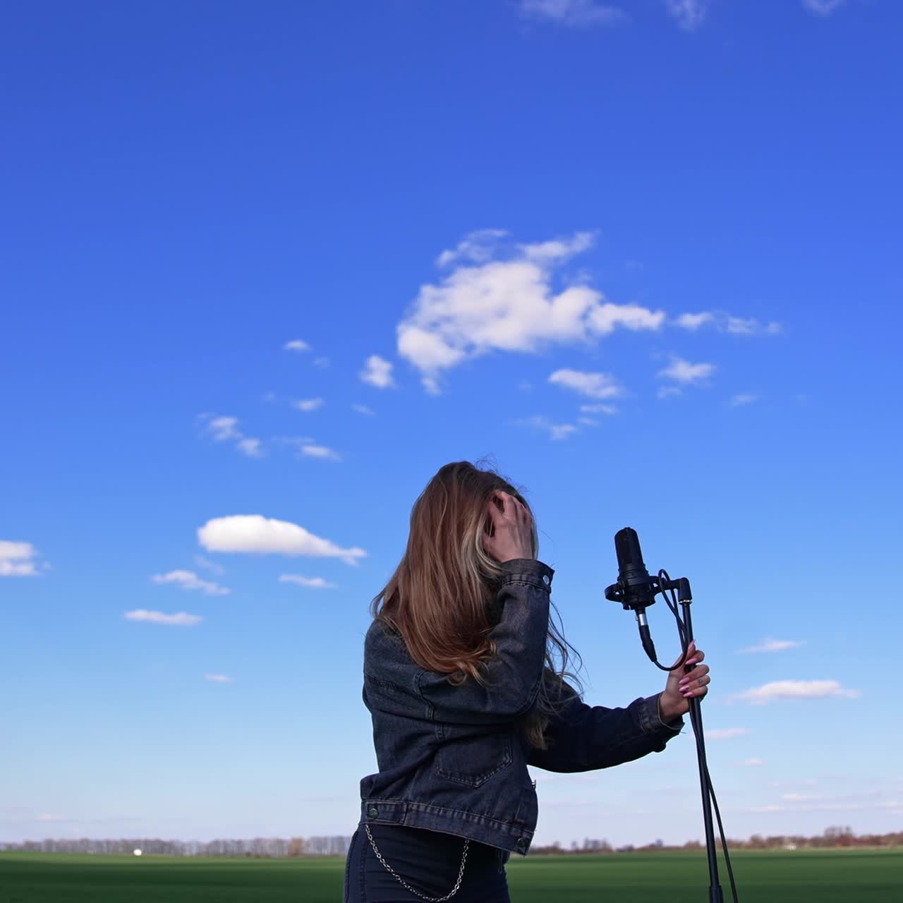 Portrait of a happy female singer on field. Young beautiful woman in denim suit with long hair standing next to the microphone on blue sky background