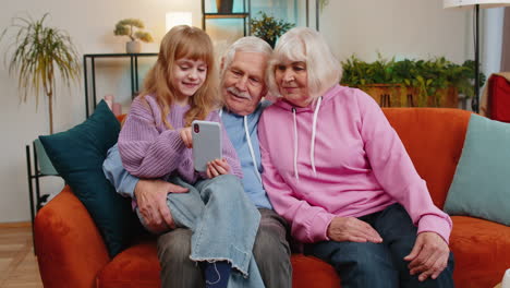 child girl granddaughter teaching grandfather and grandmother using smartphone on sofa in home room