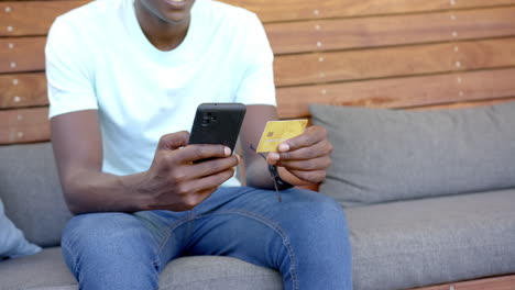 holding smartphone and credit card, african american man making online purchase on couch