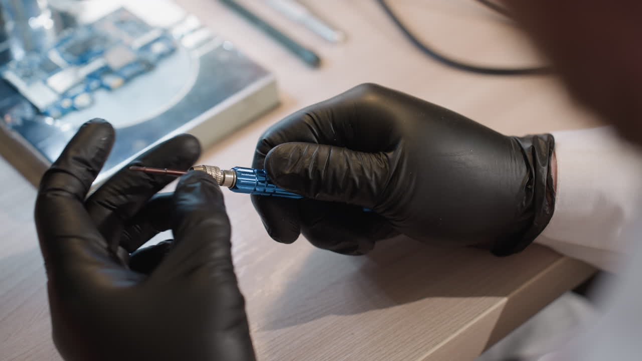 Close-up view of a technician's hands wearing black gloves, delicately turning a blue screw, with a circuit board in the background