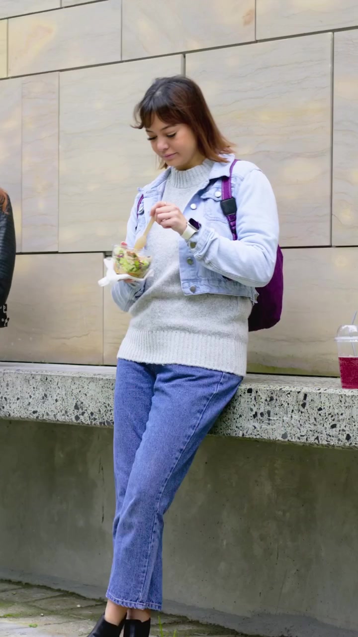 Video verticale: Donna che cammina con zaino viola e si unisce a un'amica seduta su una panchina, mangiando insalata.