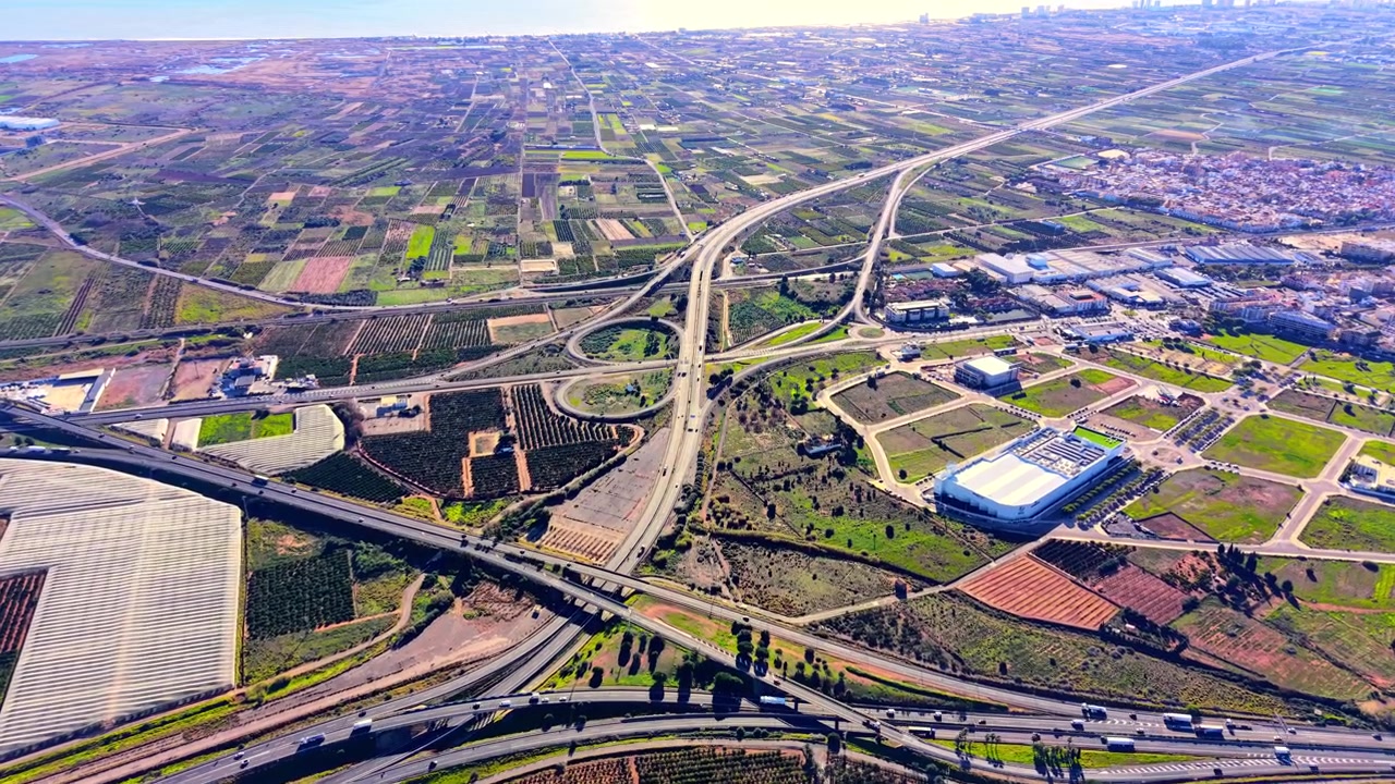 Grande entroncamento rodoviário com rampas e pontes integrado numa paisagem agrícola rural. Complexo nó rodoviário cercado por plantações agrícolas.
