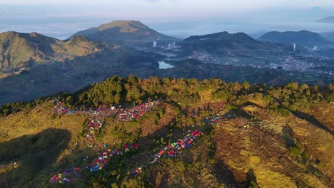Stunning aerial footage of a colorful campsite spread across a mountain ridge at sunrise with scenic highland valleys in the background. Sunrise camp point of Mount Prau, Indonesia