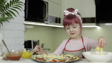 cooking. little child in apron adding grated cheese to raw pizza in kitchen