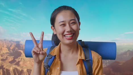 close up of asian female hiker with mountaineering backpack smiling and showing peace gesture while traveling at the top of mountain