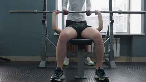 female client lying on bench lifting barbell with support from male trainer during gym workout session, focus on teamwork and fitness effort with natural outdoor daylight visible through large window