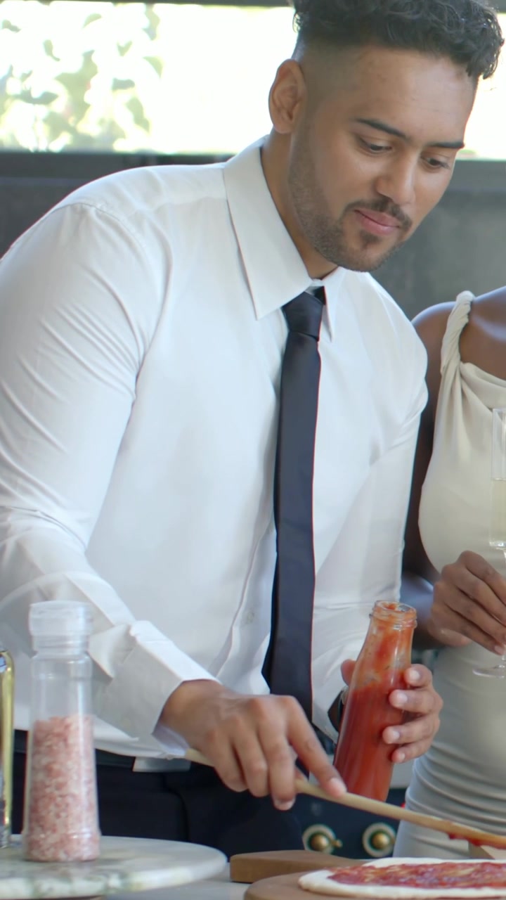 Video vertical: Exprimiendo una botella, hombre con corbata untando salsa en una pizza en el mostrador con una cuchara de madera.