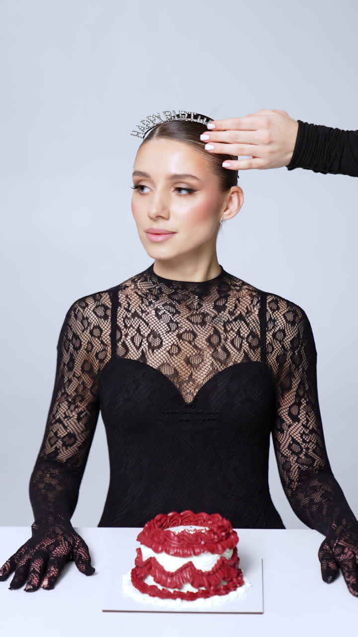 Woman in black lace outfit sits at the table with a cake. Female hands put on diadem on model's head and male hands put on sunglasses with birthday girl signs. Lady gets a lit candle, puts it into a cake and blows it. Vertical video.