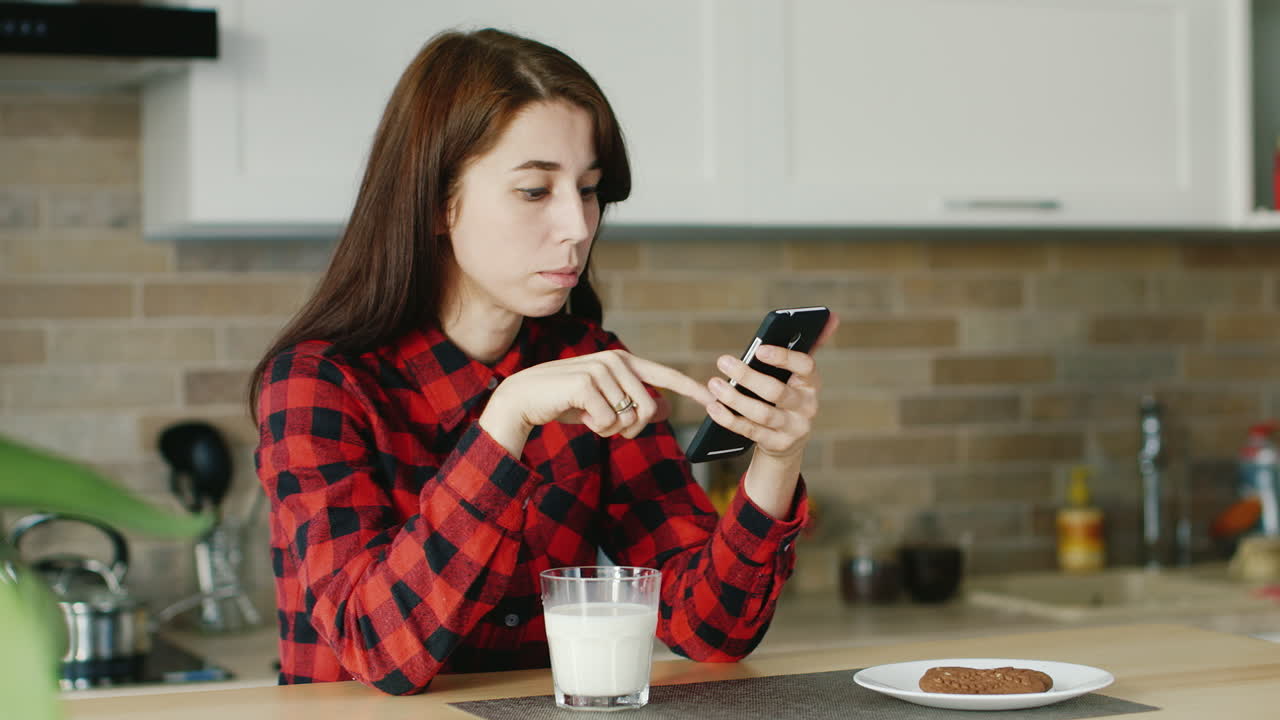 una mujer joven usa un teléfono inteligente que está sentado en su cocina junto a un vaso de salud con leche