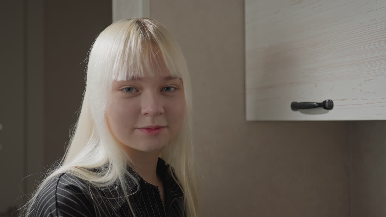 close up of blonde woman in striped shirt turning from kitchen cabinet tasks to camera with gentle smile soft light highlights sleek hair kitchen setting creates welcoming lifestyle