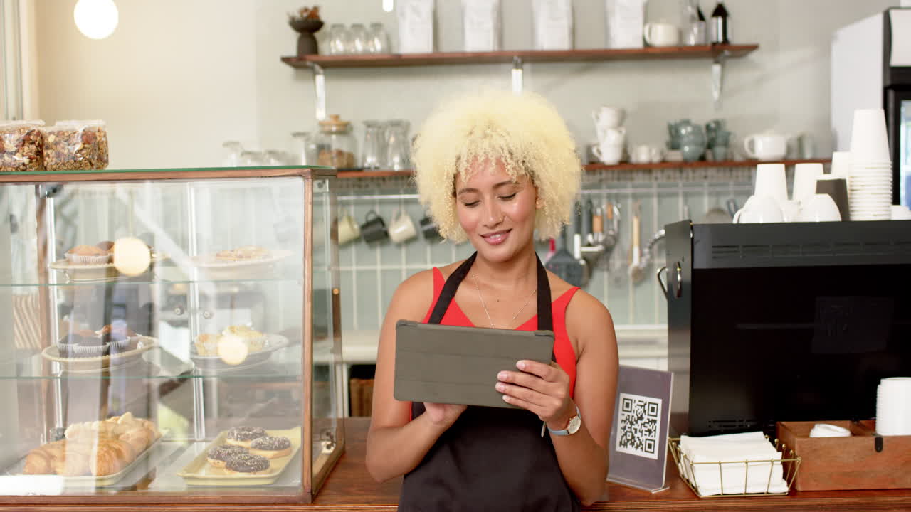 Young biracial female barista with curly blonde hair holds a tablet in a bakery