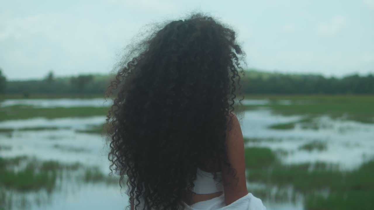 Une femme aux longs et épais cheveux bouclés de type 3C et à la peau brun foncé fait face à un marais inondé sous une douce lumière naturelle, ses cheveux doucement soulevés par le vent, évoquant le calme et l'introspection.