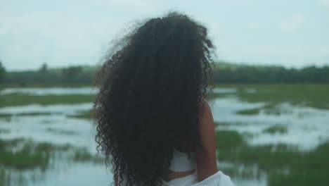 a woman with long, thick type 3c curls and dark brown skin faces a flooded marshland under soft natural light, her hair gently lifted by the wind, evoking calm and introspection