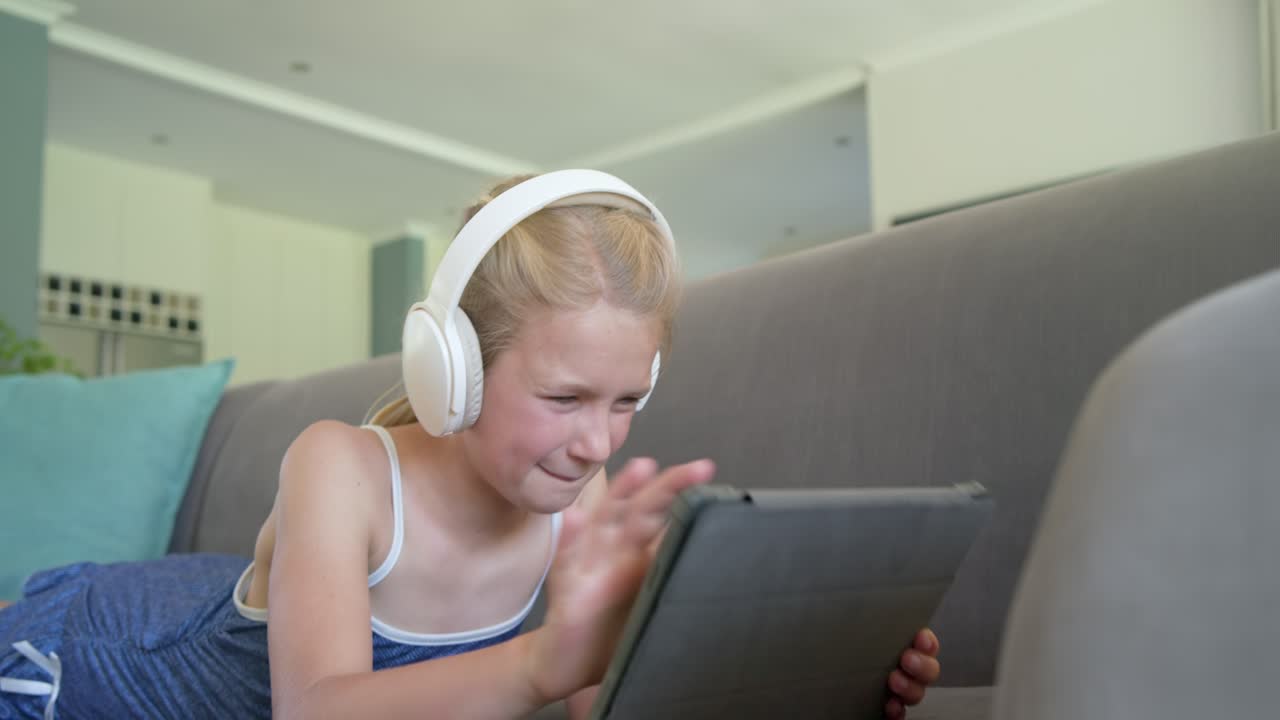 Ragazza preadolescente su divano grigio, che indossa un top blu e cuffie bianche, che risponde all'audio del tablet, toccando lo schermo