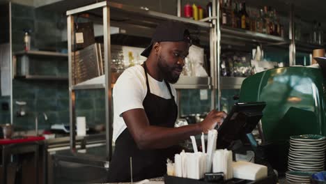 Stylish-Black-person-in-a-cap---an-employee-of-the-doner-market-takes-an-order-and-works-on-an-electronic-guy-in-a-cafe