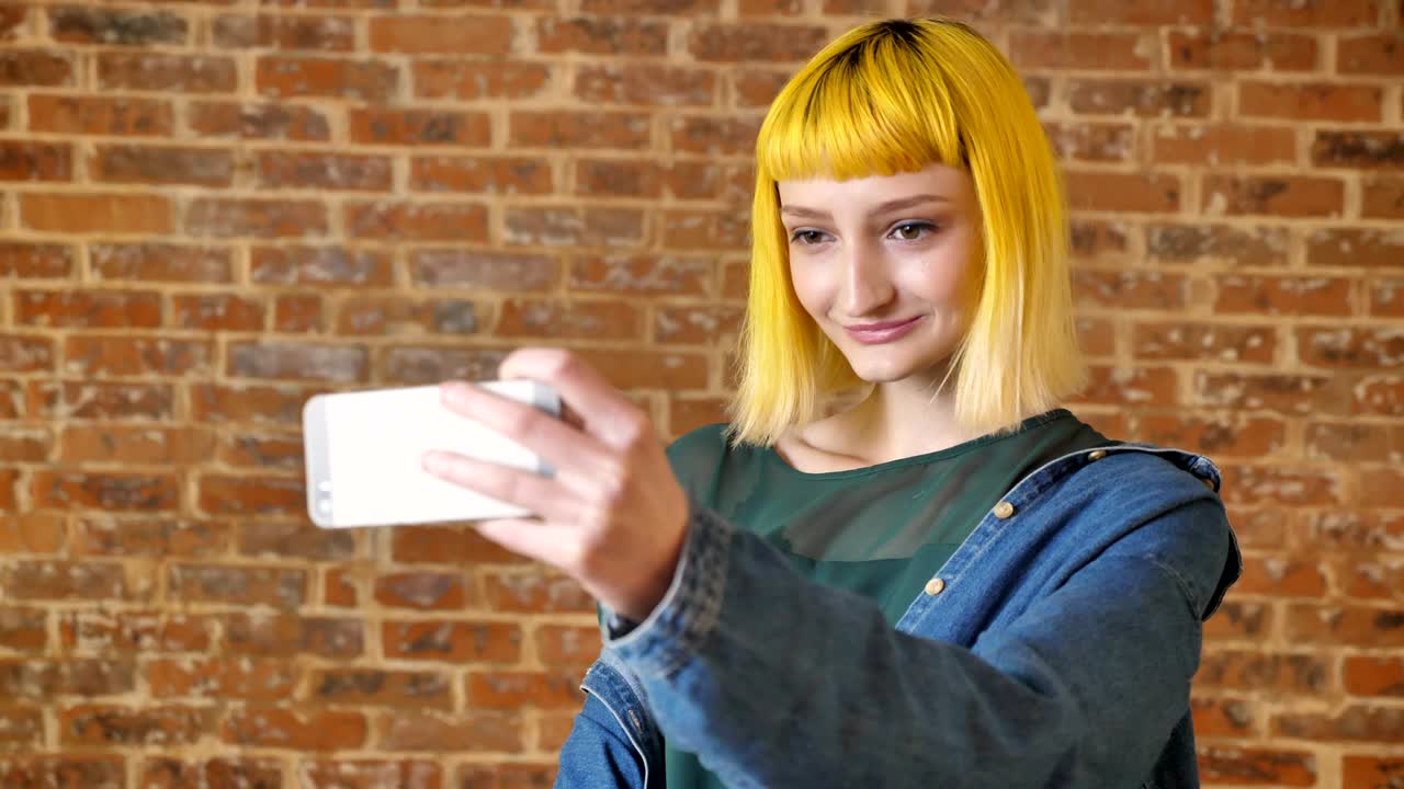 joven mujer encantadora con cabello amarillo tomando selfie con el teléfono, sonriendo y de pie cerca de la pared de ladrillo fondo, juventud moderna