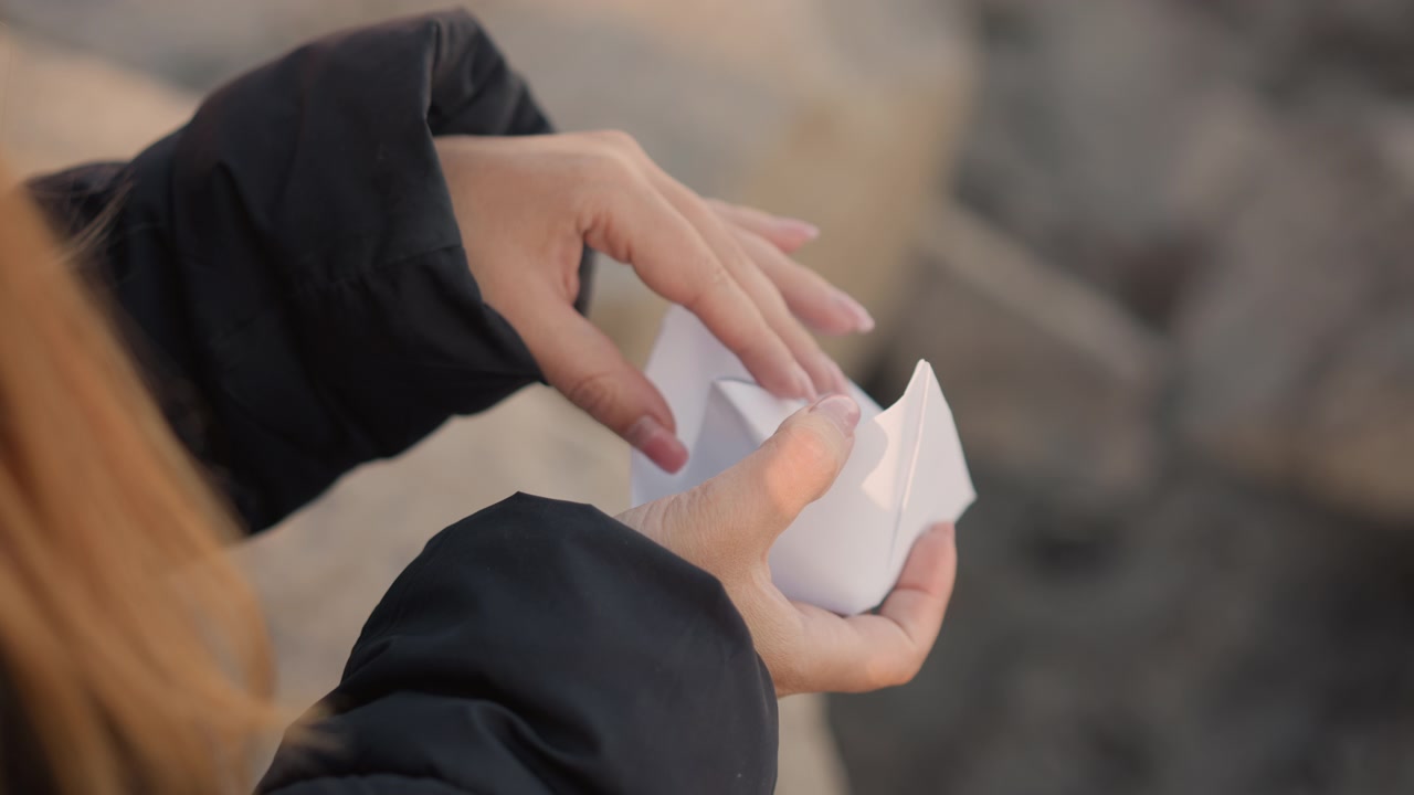 Una donna piega con cura una barchetta di carta bianca, con le mani e l'oggetto sempre a fuoco. La donna indossa una giacca nera mentre manipola il materiale creando pieghe precise contro uno sfondo esterno.