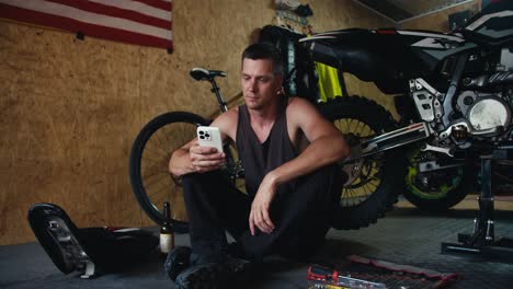 male mechanic with a short haircut in a gray t-shirt works on his white phone in the workshop during a break while repairing a motorcycle. a man sits near the tools in his workshop