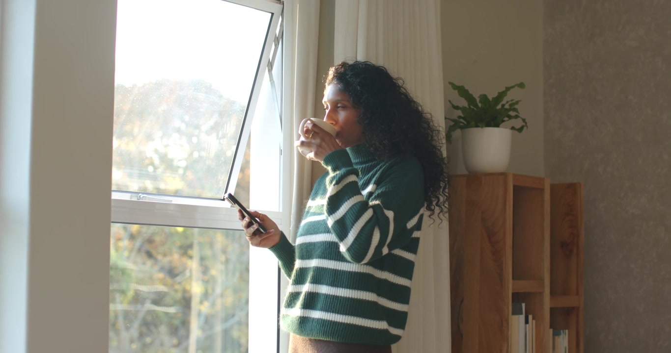 Il sole mattutino illumina una donna con un maglione verde alla finestra aperta, che sorseggia da una tazza e controlla lo smartphone.