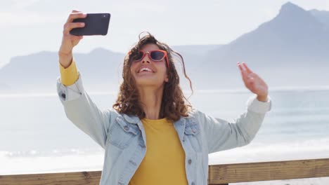 happy mixed race woman taking selfie and smiling with arm in the air on sunny promenade by the sea