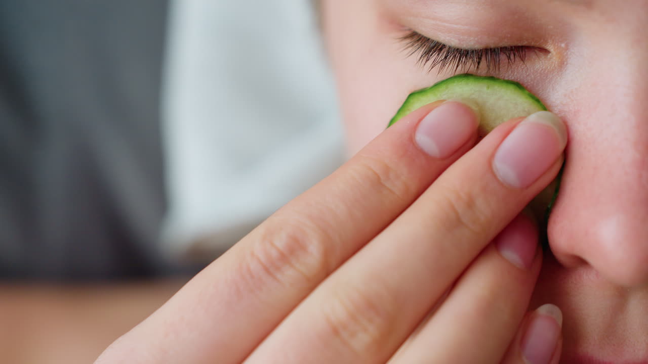 Close-up side view of woman placing cucumber slice over her eye for relaxation, emphasizes soothing self-care routine with cucumber slice applied gently to her skin for cooling