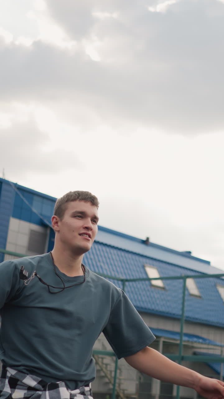 atleta con silbato alrededor del cuello lanza la pelota de voleibol hacia arriba, salta y lo golpea, cancha de baloncesto detrás con edificio azul y arena deportiva visible en el fondo, momento atlético lleno de acción