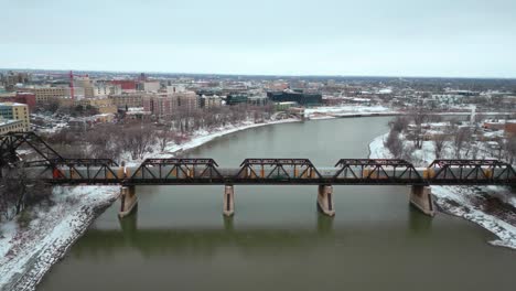 Breiter-4K-Winterschnee-Bedeckte-Eiskalte-Nördliche-Landschaft-Drohnenaufnahme-Eisen-Stahl-Zug-Industriefahrzeug-Brücke-über-Den-Assiniboine-Red-River-Grünes-Wasser-In-Winnipeg,-Manitoba,-Kanada