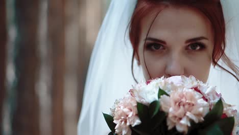 bride-stands-near-trees-in-the-forest-brings-a-bouquet-of-flowers-to-face-and-looks-at-the-camera-portrait-close-up