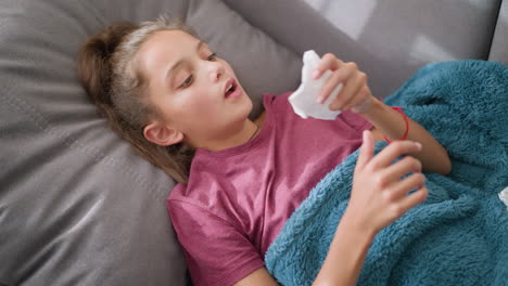 aerial view of sick child lying on couch covered with soft blue blanket, holding tissue in left hand with red bangle, sneezing and resting, showing symptoms of cold or flu, feeling unwell