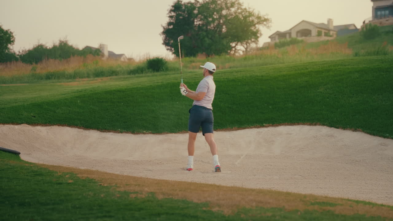 A male golfer hits out of a fairway bunker in slow motion. Sand flies as the club makes contact. The moment captures athletic precision and power during a tough shot on a bright golf course.