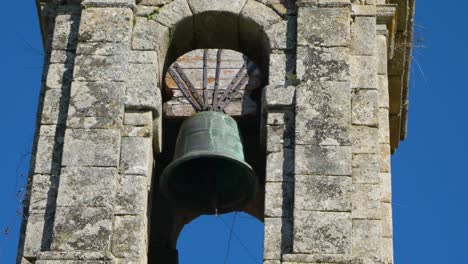 Pan-across-church-bell-suspended-in-old-weathered-stone-cut-tower-fronting-blue-sky