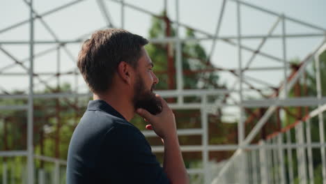 young man touching beard thoughtfully while looking into distance beside metal frame structure rural setting sunlight casting soft shadows on face and shirt