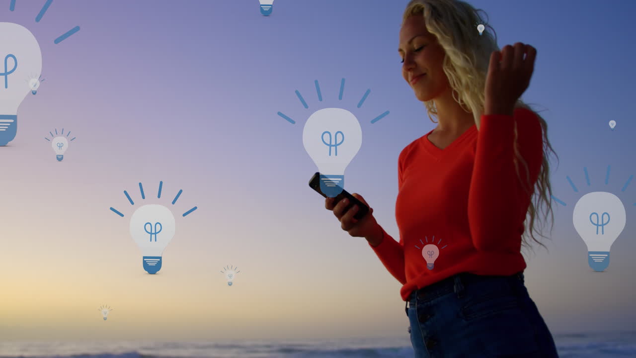 Animation of bulb icons over midsection of caucasian woman using smartphone at sunset