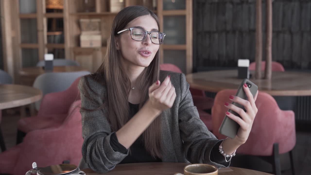 hermosa chica con anteojos sentada en un café, usando su teléfono inteligente y tomando selfies divertidos