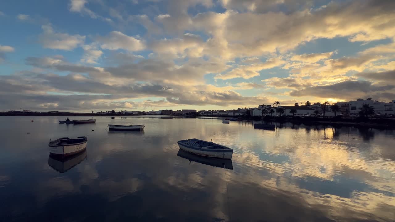 스페인 바르바테의 조용한 바다에서 휴식을 취하는 작은 시 보트들의 조용한 장면, 해가 뜨면, 물 표면에 반사되는 다채로운 구름과 배경에 보이는 마을