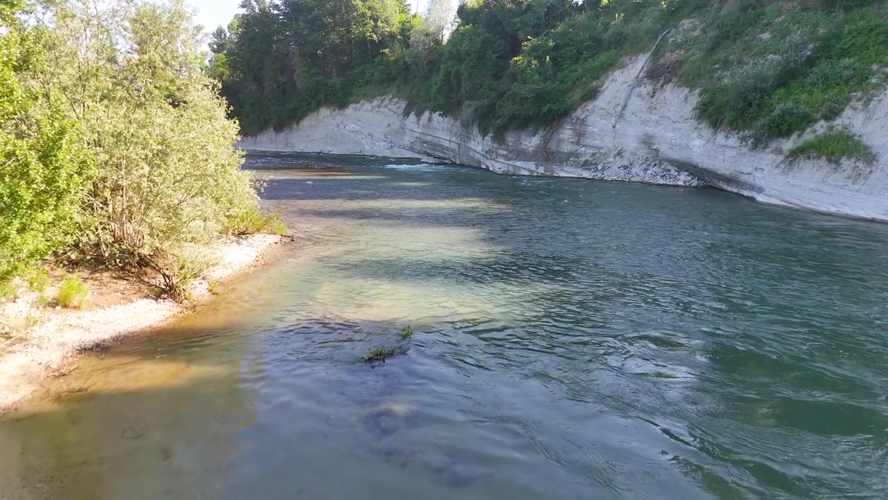Inquadratura ampia e statica di un fiume che scorre tra alberi verdi e scogliere rocciose bianche.