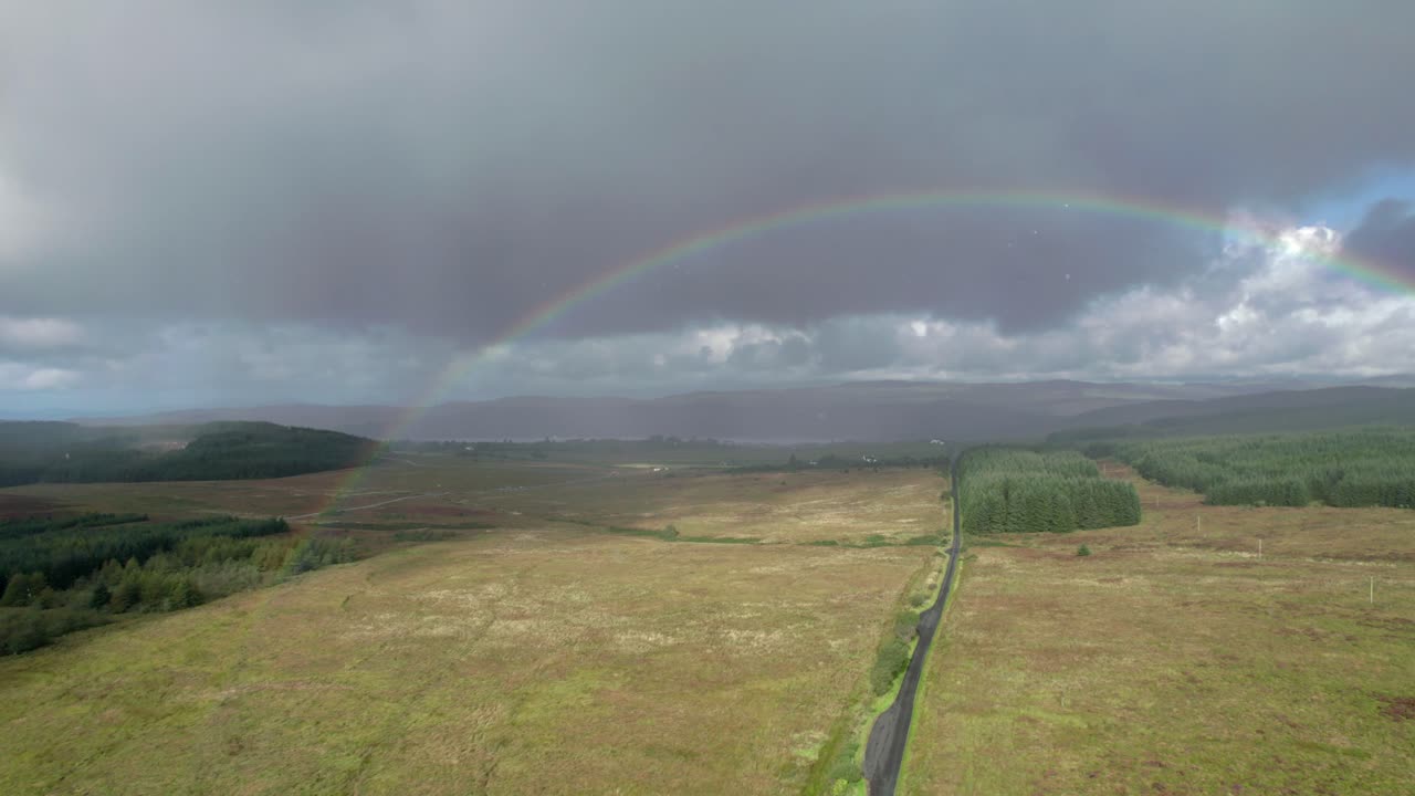 imágenes aéreas de drones que descienden bajo la lluvia sobre un camino largo y recto que mira hacia un arco iris brillante con nubes grises oscuras en el fondo, con campos, árboles y páramos en escocia
