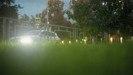close up of lush green grass with blurred view of metal railing, traffic lights, and passing car with headlamps on in vibrant urban setting during evening commute with faint trees in background