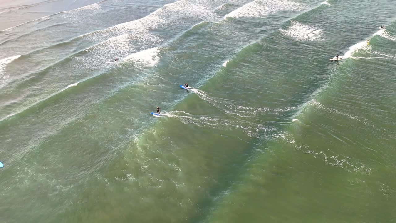 Aerial footage captures multiple surfers paddling and catching small waves in clear, shallow water under soft natural daylight at West Sands Beach